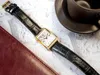 A rectangular gold-toned watch with a black leather strap is placed on a textured white surface next to a dark red notebook and a dark brown coffee cup. Produktbild-Vorschau