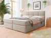 A bedroom scene featuring a boxspring bed with a headboard, mattress, and topper. The bed is dressed with pillows and a throw blanket. A large potted plant is visible to the left, and a small round side table with books is to the right. The room has large  Náhled obrázku produktu