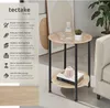 A round, two-tiered side table with a wood-look top and black metal legs, placed in a living room setting next to a sofa. The table has a bottle of water, a glass, and a small plant on the top tier, and books and a decorative item on the bottom tier. A pan Náhled obrázku produktu