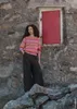 A woman stands in front of a stone wall with a red door, wearing a striped sweater and wide-leg pants. Produktbild-Vorschau