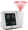 A white Braun projection alarm clock is shown against a white background. The clock displays the time 12:15 AM and an alarm time of 7:00 AM. A red projection beam emanates from the top of the clock, displaying numbers on an unseen surface. Produktbild-Vorschau