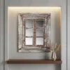 A decorative mirror designed to look like a window with shutters, placed on a wall above a shelf with a vase of dried flowers. Náhled obrázku produktu
