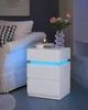 A white LED nightstand with three drawers is placed next to a sofa in a bedroom. Produktbild-Vorschau