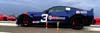 A blue race car with red accents and racing decals is parked on a track, with two red cars in the background. There are two trophies in front of the blue car. Produktbild-Vorschau