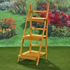 A wooden plant stand with three shelves is placed in a garden setting with flowers and greenery in the background. Produktbild-Vorschau