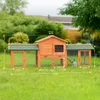 A wooden rabbit hutch with green roof, set up in a grassy backyard with trees and a house in the background. Dimensions are marked on the hutch. Produktbild-Vorschau