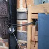 A black cable lock is wrapped around a black pole and a wooden chair leg, with a brick wall in the background. Podgląd zdjęcia produktu
