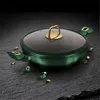 A green pot with a glass lid and gold accents, placed on a dark, textured surface with scattered gold and green gems. Anteprima dell’immagine prodotto