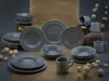 A collection of grey ceramic dinnerware, including plates, bowls, and mugs, arranged on a textured gold and grey surface. Náhľad obrázku produktu