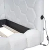 Close-up of the headboard and frame of a white upholstered bed. The headboard has a hexagonal quilted pattern. A black power cord with an adapter is visible, plugged into a USB port on the side of the headboard. The wooden slats of the bed frame are also v Náhled obrázku produktu