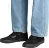 A close-up view of a person's feet wearing black Vans sneakers and light blue jeans. Voir les images du produit