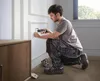 A man is kneeling and using a cordless screwdriver to install an electrical outlet on a white wall. A wooden cabinet is to the left, and a toolbox is on the floor. Voir les images du produit