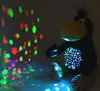 A plush toy raven is sitting in the dark, projecting colorful star and circle shapes onto a wall. Produktbild-Vorschau