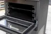 Close-up view of the interior of a black wood-burning stove with its door open, showing the oven compartment and racks. Produktbild-Vorschau