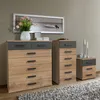A bedroom scene with three pieces of furniture: a large chest of drawers, a tall chest of drawers, and a nightstand, all with a wood and graphite finish. A framed picture of a boat hangs on the wall above the furniture, and a vase with flowers sits on top  Produktbild-Vorschau