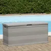 A grey storage box is placed on a brick patio next to a swimming pool with a green hedge in the background. Produktbild-Vorschau