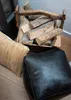 A black leather pouf and a woven pillow are placed on a wooden floor next to a rustic wooden crate filled with firewood. Produktbild-Vorschau