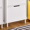 Close-up of a white shoe cabinet with ribbed doors and thin legs, placed on a wooden floor with a striped rug. Produktbild-Vorschau