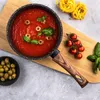 A cooking pot filled with tomato sauce, garnished with basil and olives, sits on a wooden cutting board next to cherry tomatoes and pasta. Produktbild-Vorschau