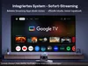 A large television screen displaying the Google TV interface, showcasing various streaming app logos and content. A small projector is placed on a stand in front of the TV. Produktbild-Vorschau