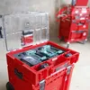 A red tool case with a clear lid, containing green power tools and batteries, is shown in a workshop or storage area. Another red tool cart is visible in the background. Náhled obrázku produktu