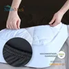 A person's hands are shown tucking a black fitted sheet onto a white mattress. A close-up inset shows the elasticized edge of the sheet. Text and logos are present. Produktbild-Vorschau