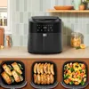 A black air fryer is shown on a kitchen counter, with three separate trays of food displayed below it: salmon, sausages, and mixed vegetables. Produktbild-Vorschau