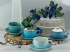 Four blue ceramic cups with saucers, two filled with drinks, are arranged on wooden slices with decorative branches and blue flowers in the background. Náhľad obrázku produktu