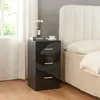 A black, high-gloss bedside table with three drawers is placed next to a bed with a white headboard and beige bedding. A window with white curtains is visible to the left. Produktbild-Vorschau