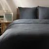 A close-up view of a bed with dark gray bedding and pillows, with a framed picture on a nightstand in the background. Produktbild-Vorschau