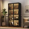 A modern living room with a black glass display cabinet filled with books and decorative items, illuminated by LED lights. A beige armchair and a potted plant are also visible. Produktbild-Vorschau