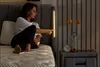 A woman is sitting on a bed and touching a light strip on the headboard. A nightstand with a clock and vase is next to the bed. Produktbild-Vorschau