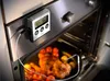 A digital meat thermometer is attached to the oven door, with its probe inserted into a roasted chicken and vegetables inside the oven. Anteprima dell’immagine prodotto