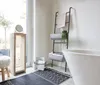 A modern bathroom with a freestanding bathtub, a ladder shelf with towels and storage baskets, a wooden stand with a mirror, and a decorative rug. Podgląd zdjęcia produktu