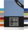 A stack of folded fabric items, likely bed sheets, with the main item in a light blue color. A black band with white text is visible on the top right corner, and a black rectangular logo with white text is at the bottom. Produktbild-Vorschau