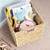 A woven storage basket filled with children's books and toys, placed on a carpeted floor next to a wall. Anteprima dell’immagine prodotto