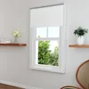 A white roller blind is installed on a window, with trees visible outside. There are shelves with decorative items on the wall. Produktbild-Vorschau