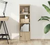 A tall, narrow bookshelf with four compartments, including a closed cabinet at the bottom, is shown against a white wall. A desk with a lamp is partially visible on the left, and a large green plant is on the right. Produktbild-Vorschau