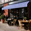 A couple sits at a table outside a cafe under a blue and white striped awning. Náhled obrázku produktu