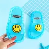 Zwei blaue durchsichtige Rutschensandalen mit gelben Smiley-Gesichtern, eine von einer Hand gehalten, auf blauem Hintergrund mit Rastermuster Náhled obrázku produktu