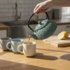 A hand pours hot water from a teal teapot into a cream mug, with two other mugs in the background. A sliced lemon rests on a wooden cutting board. Podgląd zdjęcia produktu