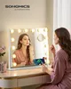 A woman applying makeup in front of a lighted vanity mirror, with a phone charging on a stand attached to the mirror. Produktbild-Vorschau