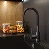 A close-up view of a modern kitchen sink area, featuring a black faucet with a flexible spout, glass jars filled with nuts, and wooden bowls. Podgląd zdjęcia produktu