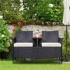 A two-seater rattan bench with a table and cushions sits on a lawn in a garden setting, with a stone wall and lush greenery in the background. Produktbild-Vorschau