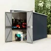 An open shed filled with tools and storage boxes, set against a textured wall and paved ground. Náhled obrázku produktu