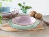 A set of colorful ceramic plates and bowls are arranged on a wooden table, with some greenery and decorative nuts in the background. Náhľad obrázku produktu