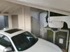 A white security camera is mounted under a carport, with a white car parked nearby. Podgląd zdjęcia produktu