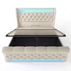 A beige upholstered bed frame with a tufted headboard and footboard, shown with the mattress support slats lifted to reveal storage space underneath. Blue LED lighting is visible along the top of the headboard and the bottom of the footboard. Anteprima dell’immagine prodotto