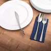 A place setting with a white plate, a knife, a spoon, and a fork on a blue napkin, all on a wooden table. Produktbild-Vorschau