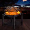 A fire pit with a mesh cover and flames burning, set on a wooden deck at dusk with a lake and silhouetted figures in the background. Produktbild-Vorschau